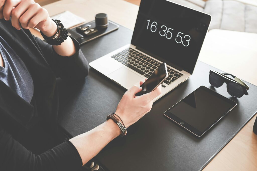 A person sitting at a desk with a laptop, phone, tablet, sunglasses, and other accessories, checking the time on a digital clock screen.